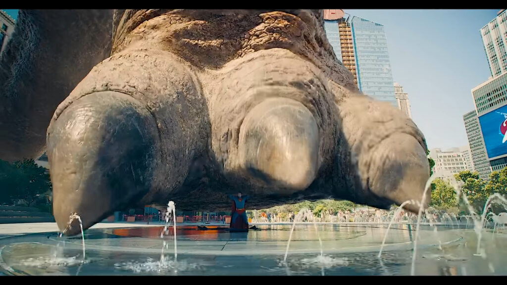 Superman braces under the massive foot of a kaiju-sized monster in a city plaza, with water fountains spraying around him.