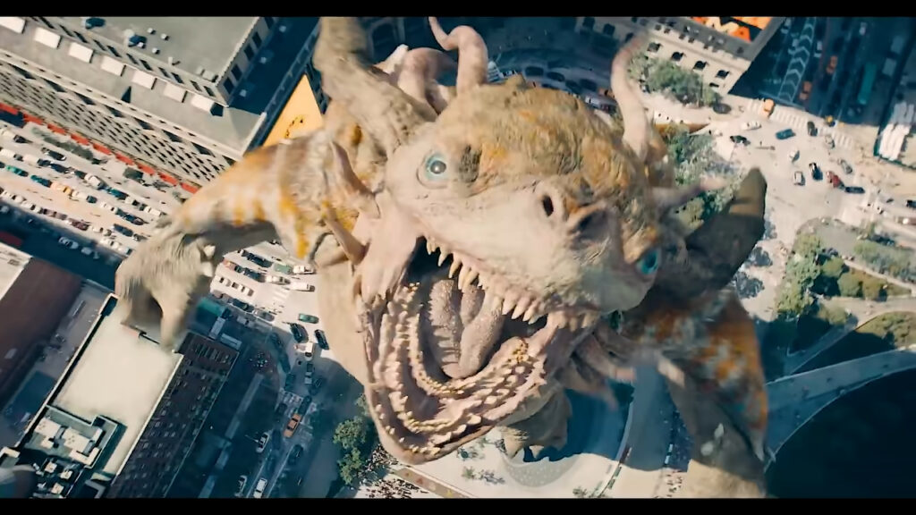 Giant kaiju roars above a city from a bird’s-eye view, revealing its massive teeth, horns, and towering size over streets and buildings.